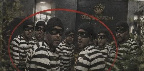 A equipe inteira, de camisa listrada de preto e branco, gorro preto e máscara nos olhos, olha surpresa para a câmera diante da garagem.