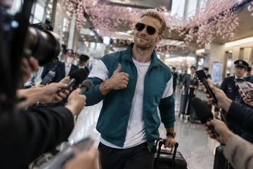 No saguão de desembarque de um aeroporto japonês, um piloto loiro de óculos escuros aponta o polegar para o próprio peito, cercado de microfones.
