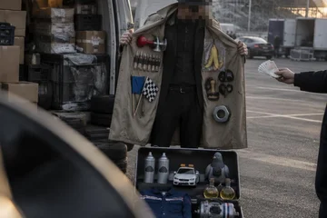 Num estacionamento, um homem de sobretudo bege, chapéu e óculos escuros abre o casaco cheio de produtos pendurados; no chão, uma mala aberta; atrás, uma van branca abarrotada.