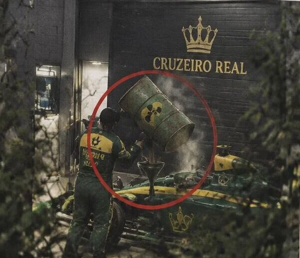 Foto granulada, tirada de longe por entre arbustos: diante da garagem da Cruzeiro Real, um homem de macacão verde e amarelo vira um tambor enferrujado com o símbolo de radioatividade num funil encaixado no carro de corrida. O tambor está circulado em vermelho.
