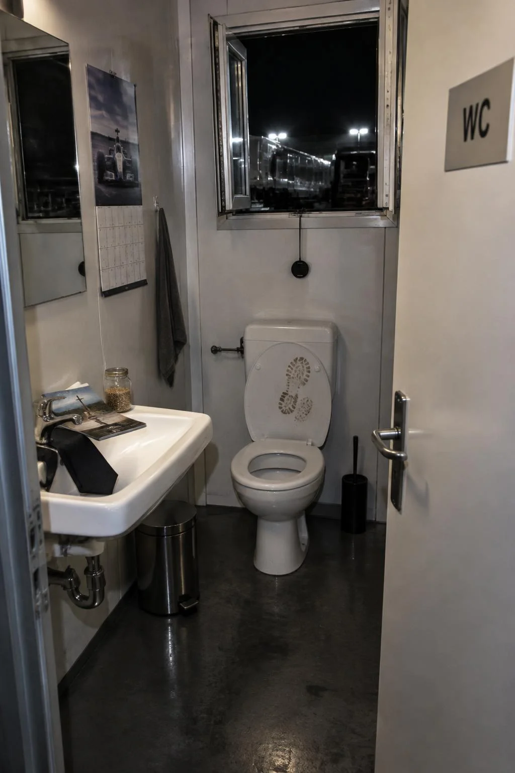 Um banheiro de funcionários vazio, com a placa WC na porta. A janelinha acima do vaso está escancarada para a noite, e há uma pegada de lama na tampa do vaso. Na pia ficaram uma gravata escura, uma revistinha religiosa e um pote de vidro com sementes de linhaça.
